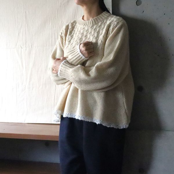 エクリュのミラノラムアンティークバスケット柄バイカラーニットに身を包み、胸の前で腕を重ねて袖口の美しい編み柄ディテールをさりげなく見せた腰上ショット。ふんわりやわらかな風合いと冬の上質感が際立つ一枚です。