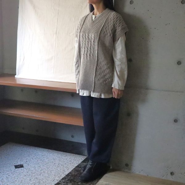 杢ベージュのNATURAL COLORED WOOL柄編みニットベストに、キナリのシャツとインディゴの裏起毛エッグパンツを合わせた女性の正面全身コーデ。自然色ウールの立体感ある編み柄が映える冬の大人ナチュラルスタイル。