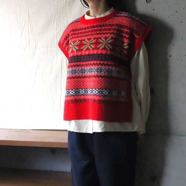 赤地に華やかなフェアアイル柄のタスマニアラムボックスベストを重ねた冬コーデの正面腰上写真。ブラウスとのレイヤードで大人かわいい温もりスタイル。