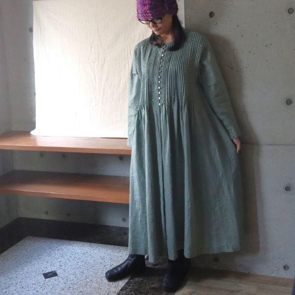 ダルグリーン色のピンタック多ボタンワンピースを一枚でさらりと着た女性の正面全身写真。落ち着いた色合いとピンタックの表情が映える大人ナチュラルなワンピースコーデ。