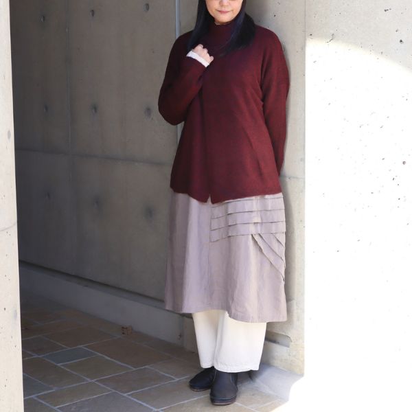裾タックデザインが印象的なアッシュグレーのワンピースの上に、ダークレッドのキャメル重ね風ニットプルオーバーを重ねた全身正面写真。裾タックがスカートのように映え、重ね着で楽しむ大人可愛いナチュラルコーデ。