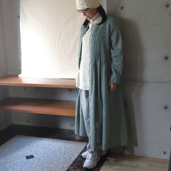 ピンタック多ボタンワンピースの前ボタンを全開し、オートミール色のタートルネックセーターと先染めヒッコリーコージーパンツを合わせて羽織った女性の正面全身写真。軽いコートのように楽しむ冬の着回しコーデ。