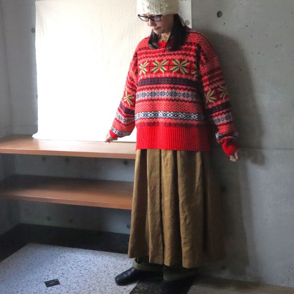 マスタード色のピンタック多ボタンワンピースに赤いフェアアイル柄セーターを重ね、オリーブ色のカツラギニューセリサイトノッポパンツを合わせた女性の正面全身写真。ワンピースをスカートのように見せた華やかで大人可愛い重ね着コーデ。