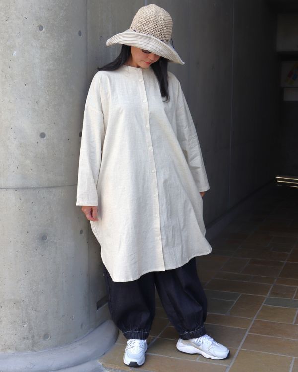 キナリ色のコットンリネンバンド襟チュニックの裾をつまみ、素材感と落ち感を見せたナチュラル系レディースコーデの全身正面写真。ロング丈チュニックとインディゴポップパンツの組み合わせ。