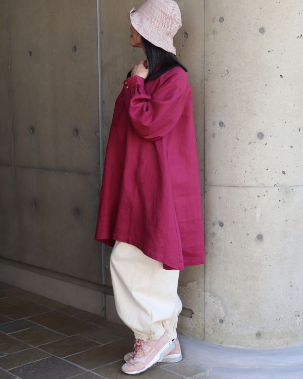 横向きで胸元に手を添え、ボリューム袖のディテールをさりげなく見せたワインレッドのヘンリーあきチュニックとキナリのポップパンツを着用したナチュラル大人女性の全身写真。